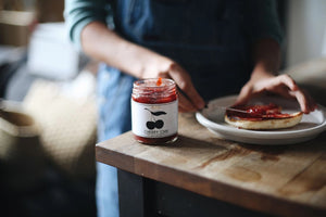 A glass jar of BRINS - CHERRY CHAI SPREAD & PRESERVE features a black lid and a label adorned with an illustration of two cherries alongside flavor details and collaboration notes. Made by BRINS with locally sourced New York sour cherries, this preserve is expertly infused with aromatic chai spices for a distinctive and flavorful experience.