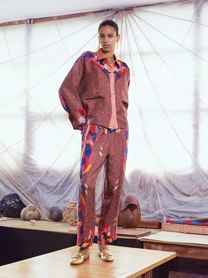 A woman stands in a well-lit room wearing FRANCO FERRARI Silk Print Trousers and a matching long-sleeve button-up top with a colorful floral design, paired with tan sandals. Her hand is in her pocket as she smiles slightly.