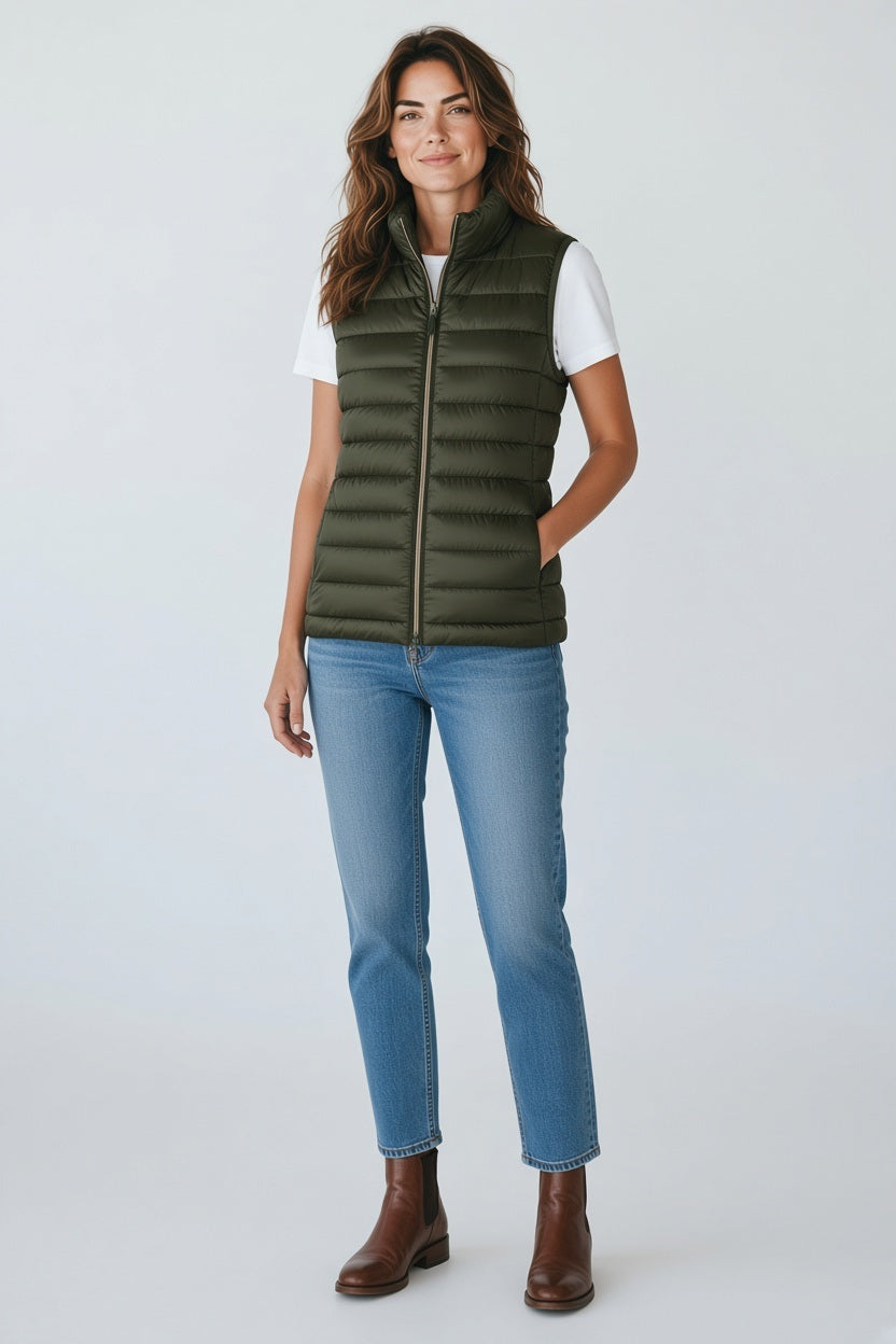 A woman stands against a plain background wearing the SAVE THE DUCK - LYNN PUFFER VEST in green, styled with a black long-sleeve top, loose black pants with pockets, and chunky black shoes. Her hands are in her pockets and she looks forward.