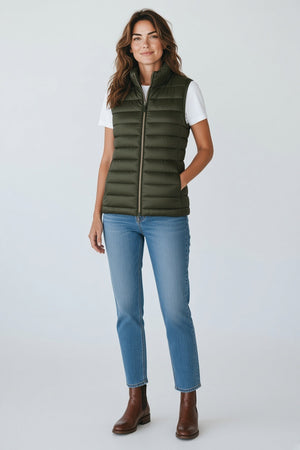 A woman stands against a plain background wearing the SAVE THE DUCK - LYNN PUFFER VEST in green, styled with a black long-sleeve top, loose black pants with pockets, and chunky black shoes. Her hands are in her pockets and she looks forward.