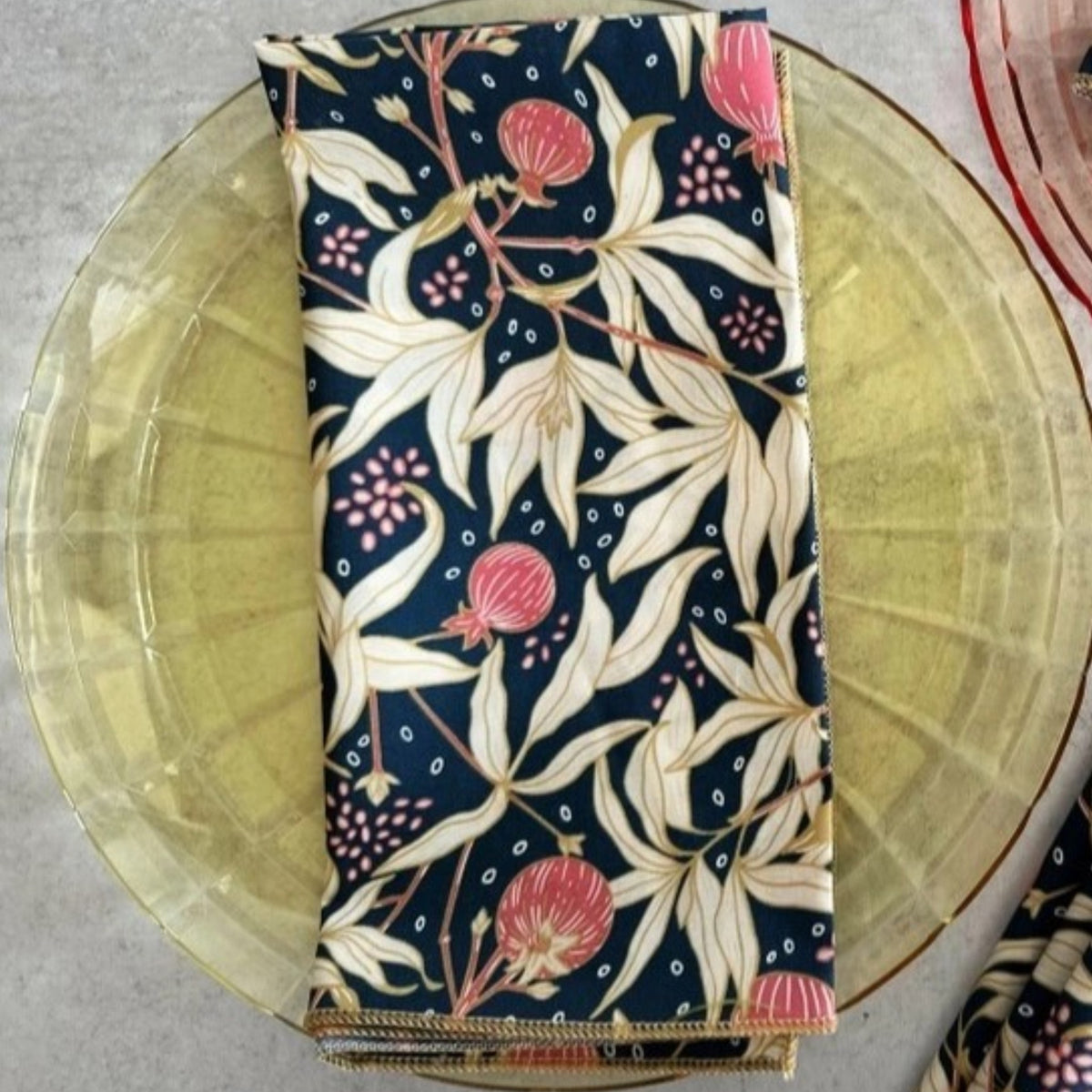 Two transparent plates, one yellow and one pink, rest on a light surface. Each displays DOT AND ARMY's Gooseberry Dinner Napkin—a dark blue cotton napkin with a beige, red, and green leaf pattern for stylish sustainable dining.