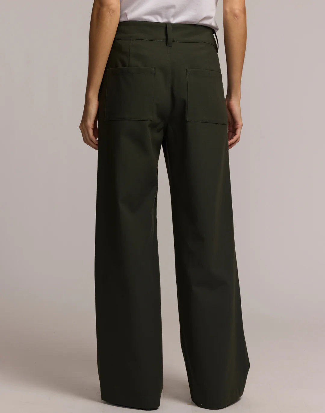 A person wears HINSON WU&#39;s KELLY TRAVELER&#39;S TWILL PANT in dark green with front pockets and gold buttons, styled with a white top and black pointed-toe shoes. Photo is cropped from shoulders down, highlighting this travel-ready look.
