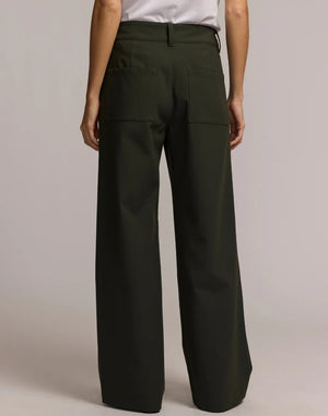 A person wears HINSON WU's KELLY TRAVELER'S TWILL PANT in dark green with front pockets and gold buttons, styled with a white top and black pointed-toe shoes. Photo is cropped from shoulders down, highlighting this travel-ready look.