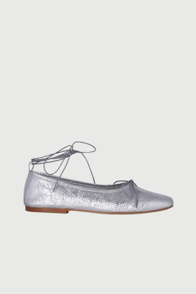The Augusta - Sienna Ballet Flat by AUGUSTA is a single silver-colored shoe featuring a subtle metallic sheen and a simple bow on the front. It has a low brown sole and a pull tab at the back. Handcrafted in Spain from leather, this ballet flat is shown from the side against a light gray background.