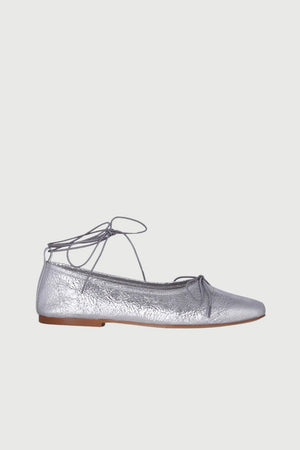 The Augusta - Sienna Ballet Flat by AUGUSTA is a single silver-colored shoe featuring a subtle metallic sheen and a simple bow on the front. It has a low brown sole and a pull tab at the back. Handcrafted in Spain from leather, this ballet flat is shown from the side against a light gray background.