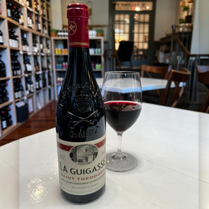 A bottle of THEODORIC LA GUIGASSE CHATEAUNEUF-DU-PAPE 2022 from WINE: A-VIN-GARDE/BORDEAUX FINE & RARE rests on a table in a wine shop, surrounded by shelves lined with assorted bottles. Also on the table are a lit candle and a small plant.