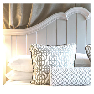 A neatly made bed with white pillows and a white textured blanket, part of the FEARRINGTON LIFESTYLE BEDDING COLLECTION - QUEEN LINEN EXPERIENCE from Fearrington House, against a light-colored, paneled wall background. The pillows slightly overlap, and soft light falls across the fabric, creating a peaceful and inviting scene.