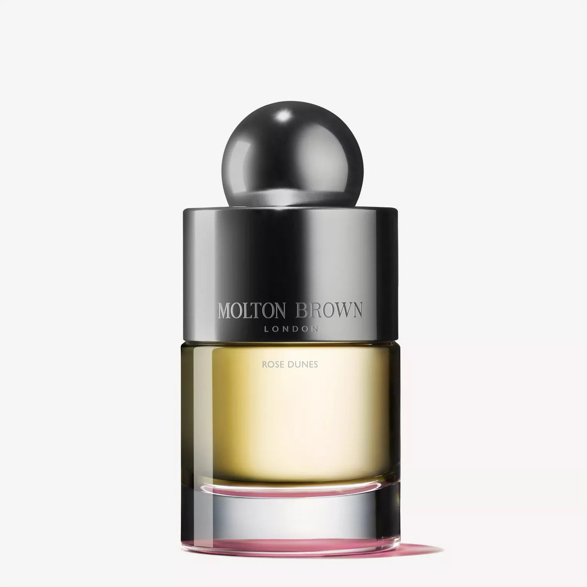 A clear glass bottle of MOLTON BROWN - ROSE DUNES EAU DE TOILETTE with a metallic cap and pink-tinted base, featuring velvety rose notes, is displayed against a plain white background.