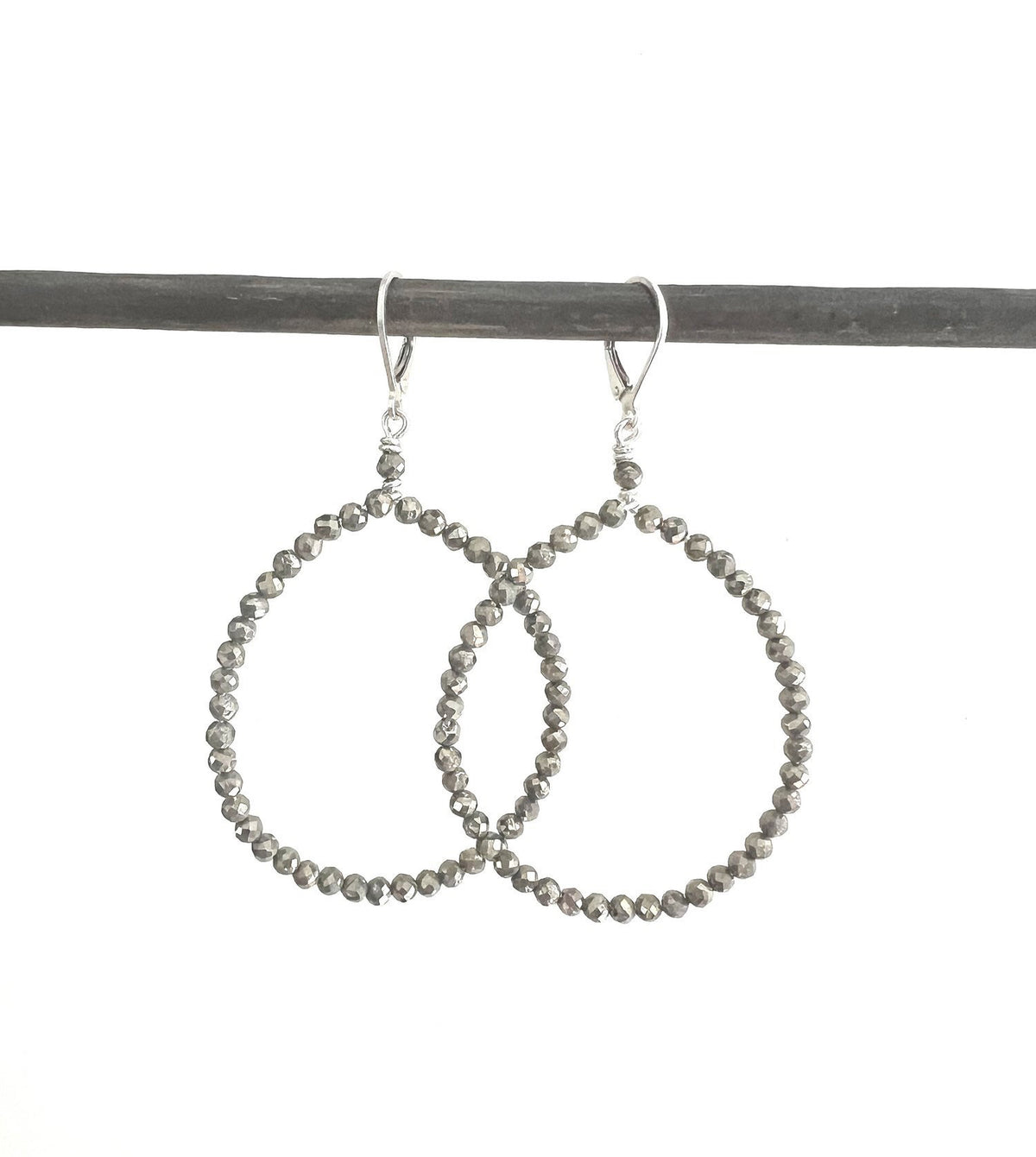 A light-skinned woman with a subtle expression gazes downward, dark hair pulled back and adorned with ANN LIGHTFOOT&#39;s Faceted Pyrite Hoop Earrings. Her soft-textured clothing contrasts against a bright, softly focused background.