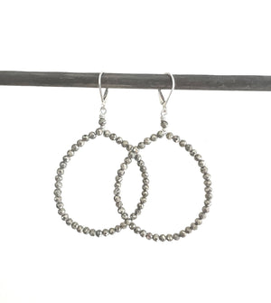 A light-skinned woman with a subtle expression gazes downward, dark hair pulled back and adorned with ANN LIGHTFOOT's Faceted Pyrite Hoop Earrings. Her soft-textured clothing contrasts against a bright, softly focused background.