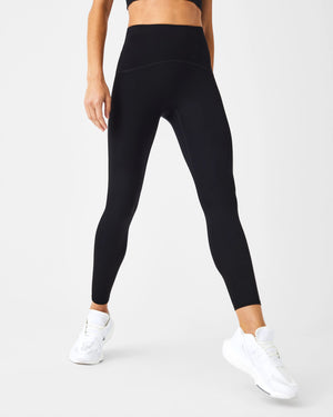 A person wears SPANX - BOOTY BOOST 7/8 LEGGINGS in high-waisted royal blue performance fabric with black athletic shoes, standing hand on hip against a plain white background. Only their lower body is visible; upper body and face are not shown.