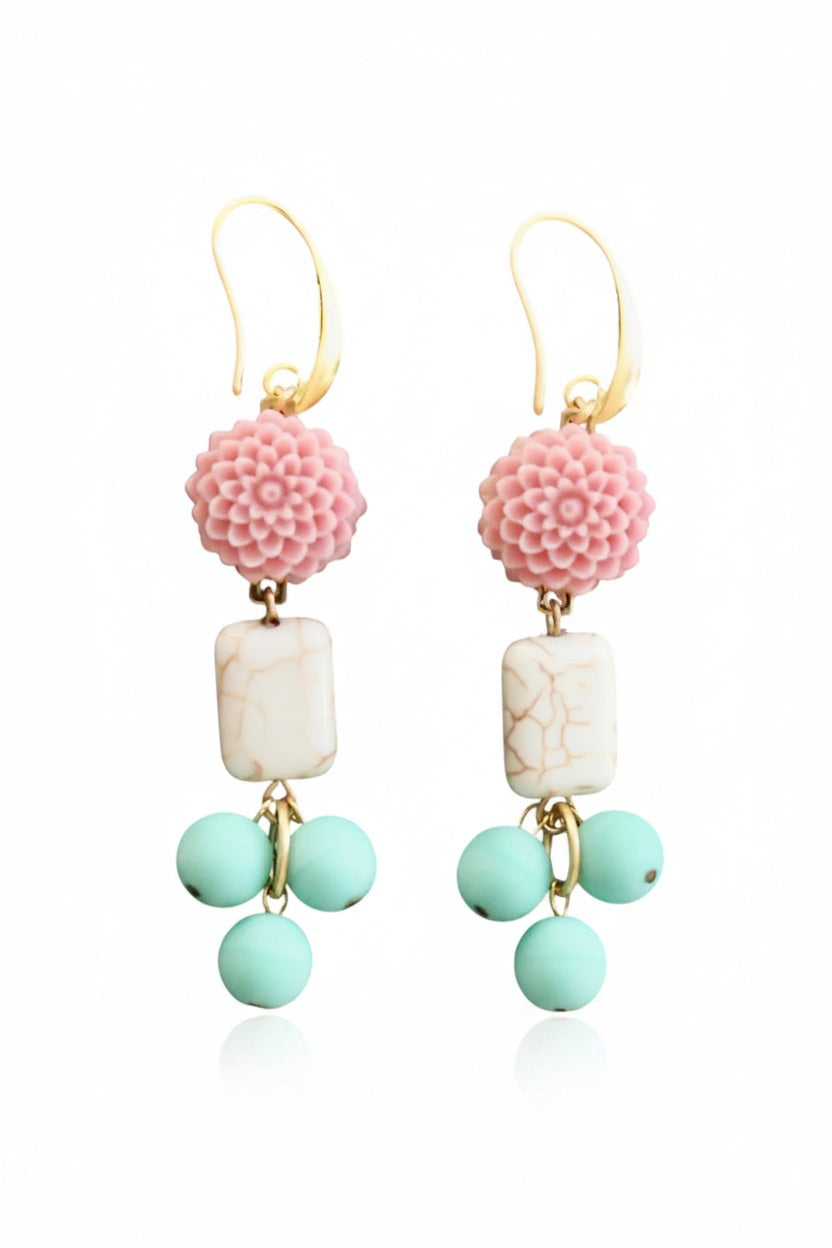 DAVID AUBREY - PINK AND TURQUOISE FLOWER EARRINGS by DAVID AUBREY feature gold hook fastenings, pink flower tops, rectangular white magnesite beads with a marble-like finish, and three dangling round mint-green Czech glass beads. Displayed on a dark hexagonal surface, these earrings blend delicate detailing with timeless elegance for a truly unique accessory.