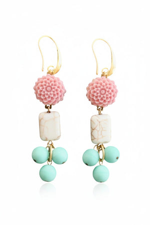 DAVID AUBREY - PINK AND TURQUOISE FLOWER EARRINGS by DAVID AUBREY feature gold hook fastenings, pink flower tops, rectangular white magnesite beads with a marble-like finish, and three dangling round mint-green Czech glass beads. Displayed on a dark hexagonal surface, these earrings blend delicate detailing with timeless elegance for a truly unique accessory.