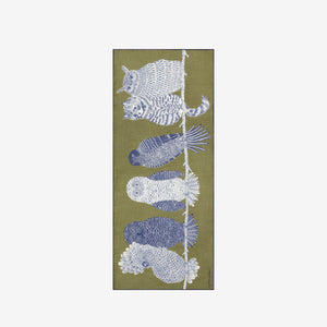 A woman with blond hair tied back wears a light gray sweater and black pants, with the INOUI EDITIONS - CHOUETTE WOOL SCARF by INOUI—an olive green wool scarf featuring a large blue and white owl design—draped over her shoulder. This hand-printed scarf, made in India, adds a vibrant touch against the plain white background.