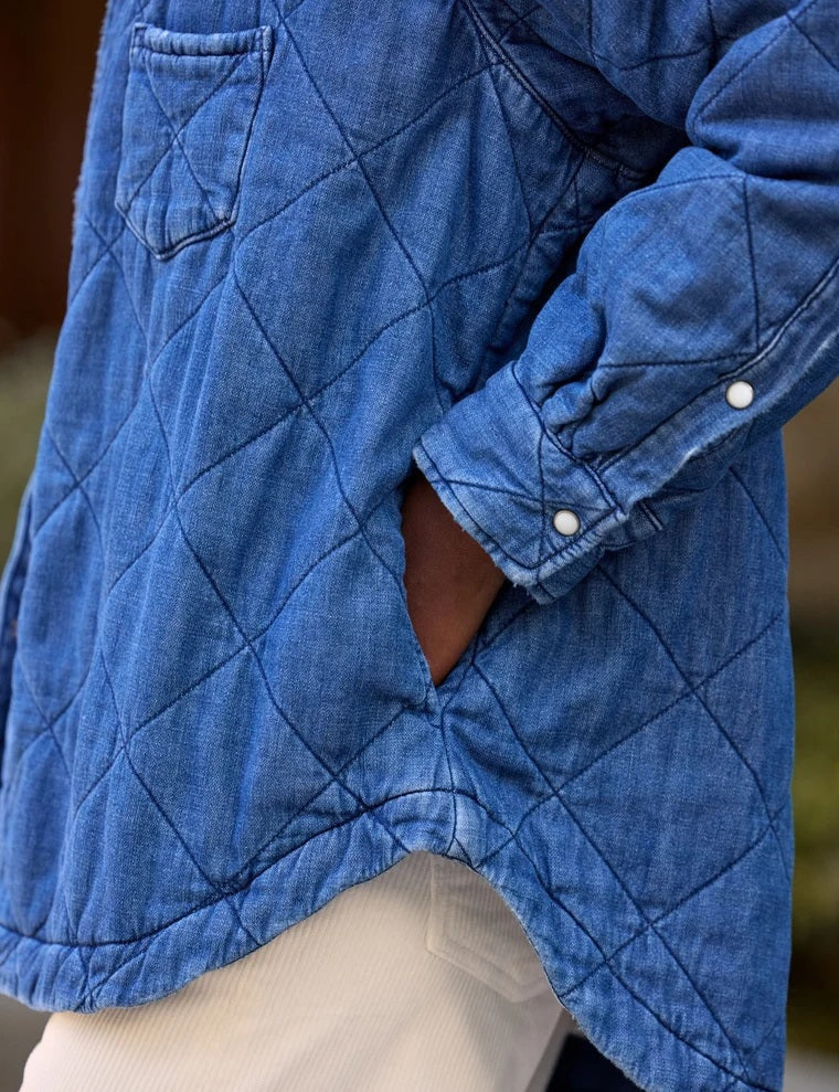 A person holds open a blue FRANK &amp; EILEEN - EILEEN Quilted Shirt Jacket in Famous Denim Distressed Indigo Wash, revealing its interior pocket and distinctive diamond quilted pattern. The person is wearing a cream-colored, ribbed top, with a blurred background.