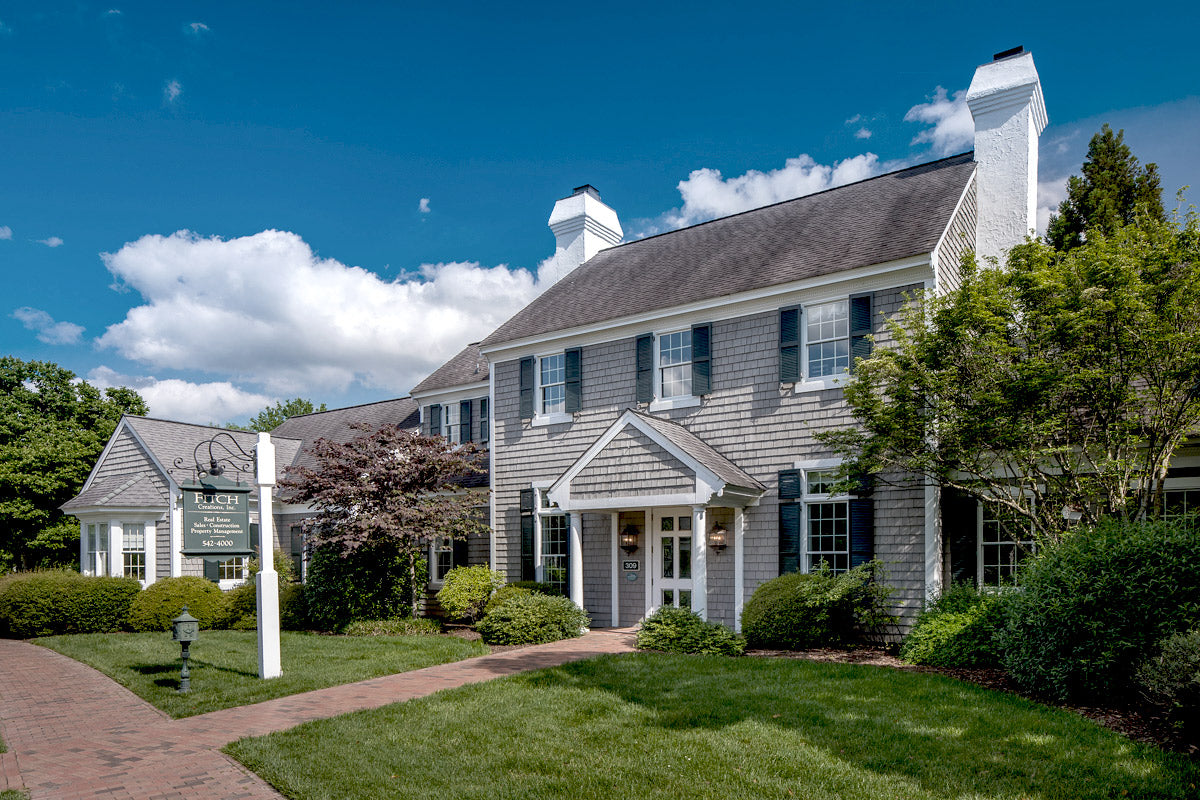 A large, gray-shingled building with white trim and two chimneys sits amid green trees and shrubs, under a blue sky with clouds. A sign near the entrance reads "Leigh Agency Insurance." Brick walkway in front.