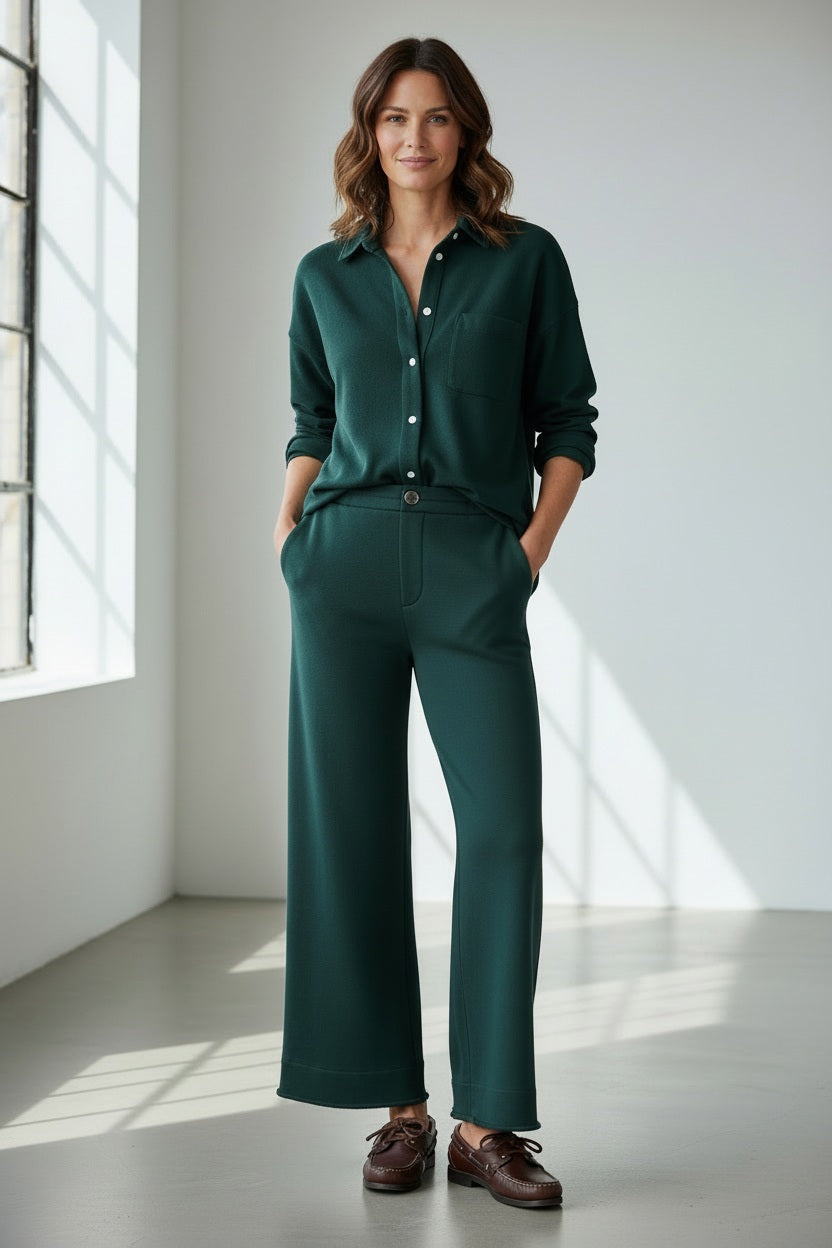 A woman stands indoors with her hands in her pockets, wearing the FRANK & EILEEN Riley Cropped Trouser Pant in Triple Fleece Pine from the California Heritage Collection, paired with a matching dark green button-up shirt and brown shoes, as sunlight streams through a large window.