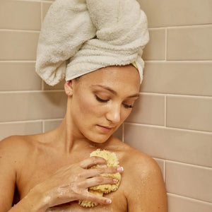A natural BAUDELAIRE bath sea sponge from the “BATH SEA SPONGE GIFT BOX, SMALL” rests beside a brown box featuring an illustrated figure and the product label. Another sustainable bath sponge from BAUDELAIRE is partially visible inside the box.