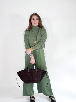A woman with long brown hair smiles against a white background, wearing a ROSE & CROWN - CASHMERE TURTLENECK and wide-legged green pants, accessorized with necklaces, bracelets, and a large black handbag.