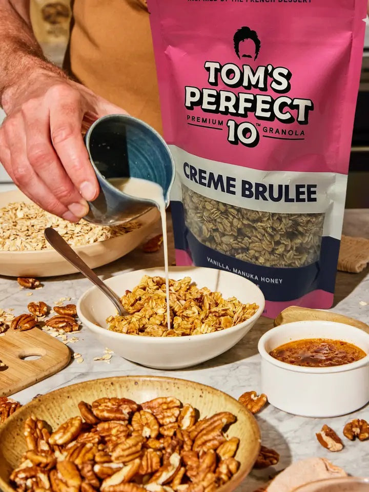 A hand pours milk over a bowl of TOM’S PERFECT 10 - CREME BRULEE GRANOLA, creating a wholesome and healthy snack. Nearby are a bag labeled “TOM’S PERFECT 10,” along with pecans, oats, manuka honey, and a small dish of crème brulee dessert on the table.