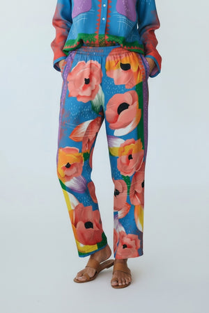 A person wears FRANCO FERRARI - SILK PRINT TROUSERS featuring a bold red, pink, and orange floral print on blue, paired with a matching top and brown sandals. Only the lower half of the body is visible.