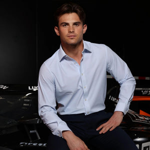 A young man with short brown hair poses against a dark background, wearing the COLLARS AND CO - APEX DRESS SHIRT SEMI SPREAD IN GLACIER BLUE TWILL by COLLARS AND CO. He gazes at the camera with a neutral expression. The partial word “LUCAS” appears to his left, evoking a sense of excellence reminiscent of the Arrow McLaren IndyCar Team.
