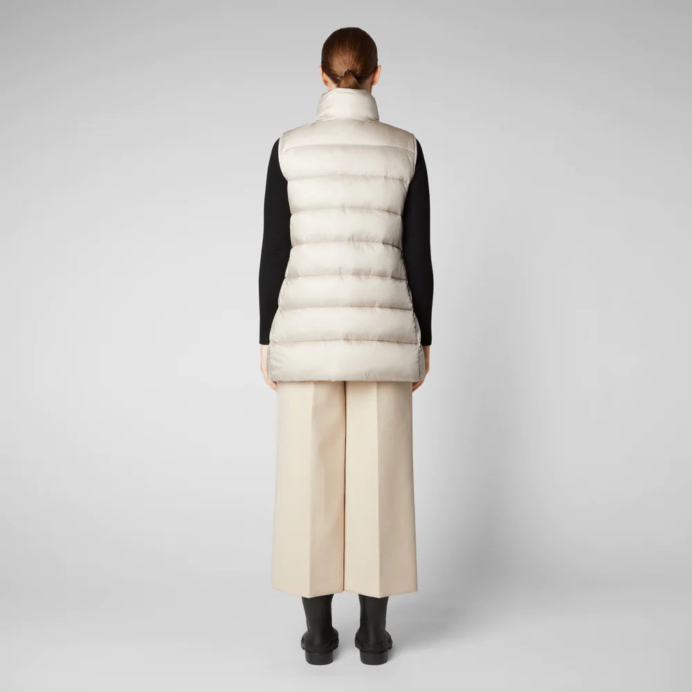 A person wearing a black cap, sunglasses, the SAVE THE DUCK - CORAL PUFFER VEST with animal-free down from SAVE THE DUCK over a black long-sleeve top, beige wide-legged pants, and black ankle boots stands against a plain background. The outfit appears to be styled for colder weather.