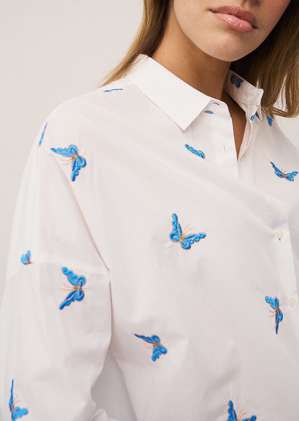 A woman with long, straight hair stands against a plain background wearing the 0039 ITALY LUCY BLOUSE WITH EMBROIDERED BUTTERFLIES, light blue wide-leg jeans, and silver sandals. She is looking slightly to the side.