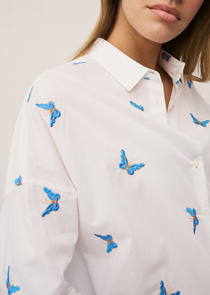 A woman with long, straight hair stands against a plain background wearing the 0039 ITALY LUCY BLOUSE WITH EMBROIDERED BUTTERFLIES, light blue wide-leg jeans, and silver sandals. She is looking slightly to the side.