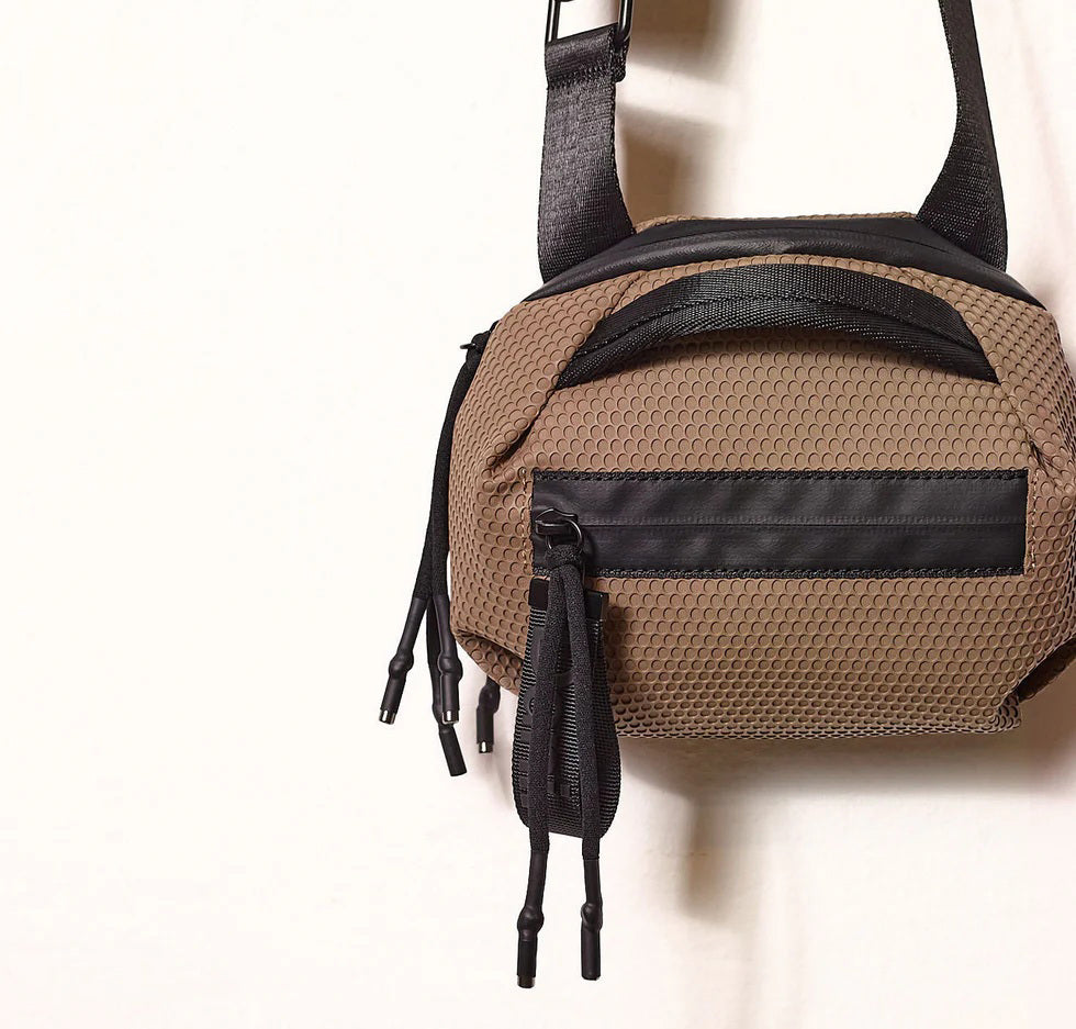 The DANIELLA LEHAVI BONNIE MINI TRAVEL CROSSBODY, a small brown vegan bag with black accents and strap, is displayed on a white pedestal against a plain beige background.