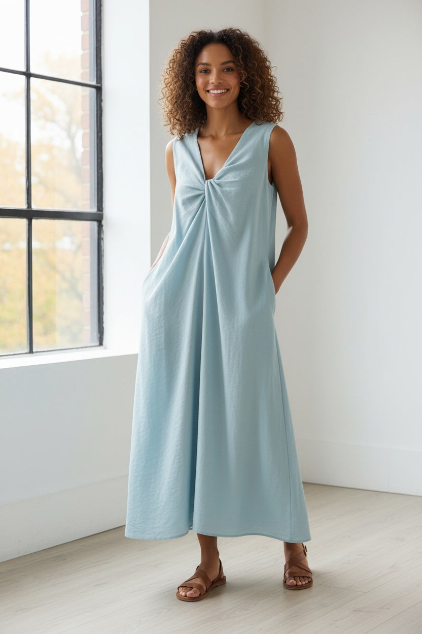 A person stands on white pebbles wearing the ELEH - DRESS WITH CENTRAL KNOT, a sleeveless sky blue A-line dress by ELEH, paired with brown sandals, against a plain white background.