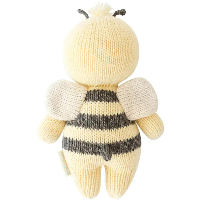 A baby lies on a cozy white blanket, raising both arms. The baby is dressed in a peach outfit and a headband with three pom-poms. Next to the baby is the CUDDLE + KIND - HANDMADE BABY BEE by CUDDLE &amp; KIND, a fair trade, hand-knit toy with black and white stripes—a perfect addition to any collection of baby shower gifts.
