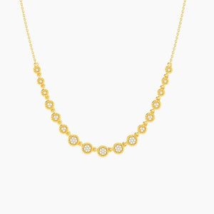 A woman with blonde hair styled in a low updo wears a white off-shoulder top and the ELLA STEIN - MULTI CIRCLE CHAIN NECKLACE by ELLA STEIN, featuring gold plated chains adorned with round, sparkling floral-shaped pendants. She looks slightly down with a calm expression against a neutral background.