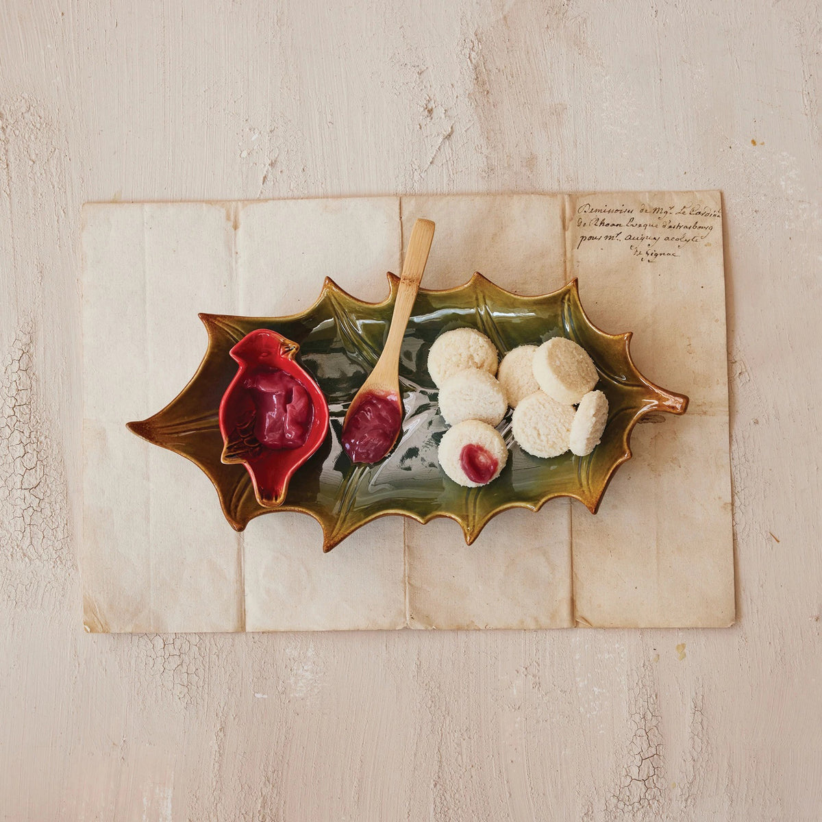 The CREATIVE COOP STONEWARE PLATE WITH CARDINAL DISH AND BAMBOO SPOON SET displays jam-filled cookies, with extra jam in the cardinal dish and a bamboo spoon, all arranged on folded paper over a light, textured surface.
