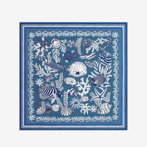 A person in a sleeveless cream top wears the INOUI EDITIONS - NEPTUNE SCARF IN BLUE by INOUI, featuring white floral and geometric patterns, draped over one shoulder against a plain white background.