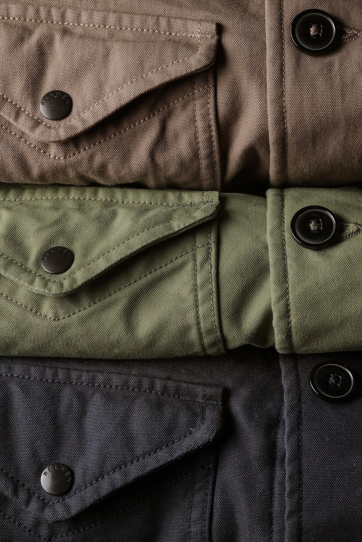 Three folded RELWEN Pile-Lined Chore Coats in Tobacco, Olive, and Charcoal are stacked, highlighting classic workwear style with buttoned front pockets and prominent stitching details. The rugged fabric texture and metal buttons of each RELWEN jacket are clearly visible.