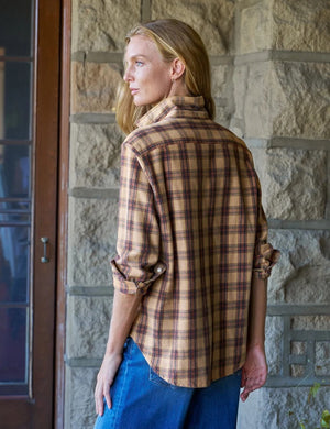 A woman stands in a sunlit room, smiling with curly hair, wearing the FRANK & EILEEN EILEEN Relaxed Button Up Shirt in Italian Flannel Gold Brown Plaid, paired with blue skinny jeans and brown ankle boots.