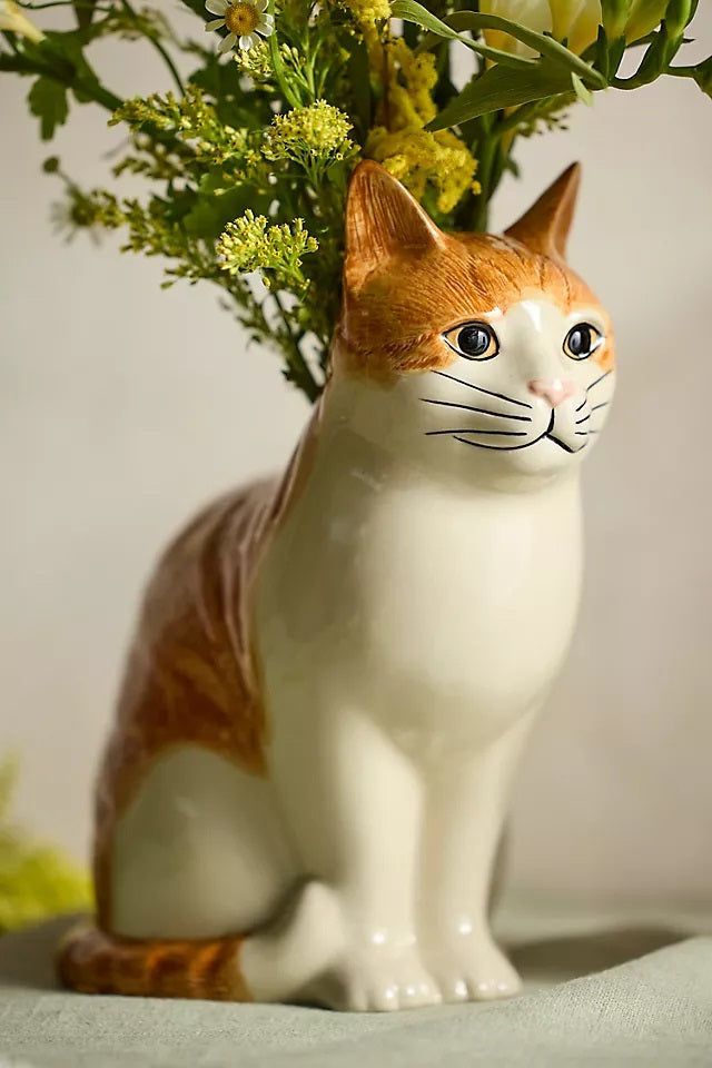 The QUAIL CERAMICS - SQUASH CAT FLOWER VASE by QUAIL DESIGNS is a hand-painted stoneware vase shaped like a sitting orange cat, with green foliage and yellow flowers appearing to grow from its back.