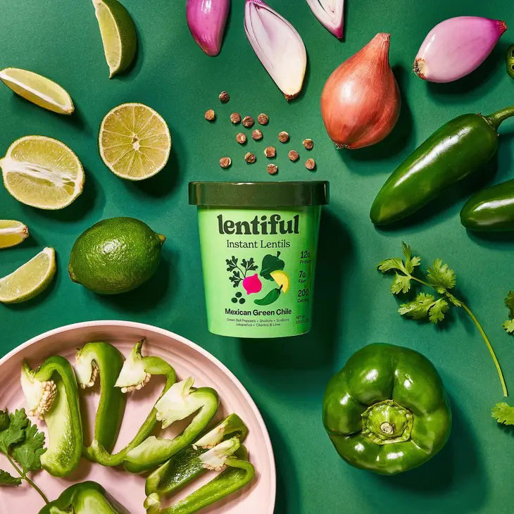 A container of LENTIFUL - MEXICAN GREEN CHILE INSTANT LENTILS is surrounded by fresh ingredients including green bell peppers, jalapeños, limes, cilantro, garlic cloves, onion slices, and green peas—all arranged on a vibrant green background.