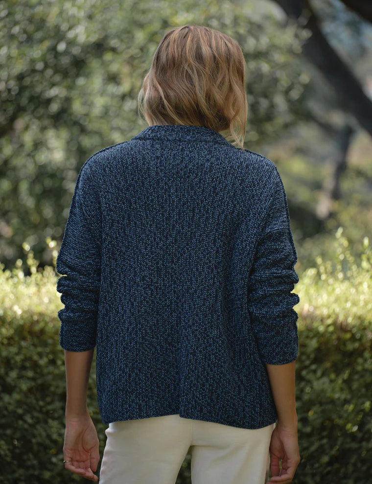 A woman stands outdoors amid lush greenery and trees, wearing the FRANK & EILEEN Montecito Crew Neck Cardigan in Pure Italian Cotton Blueberry over a white shirt and white pants, its relaxed fit perfectly complementing the natural background.