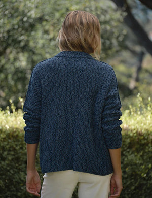 A woman stands outdoors amid lush greenery and trees, wearing the FRANK & EILEEN Montecito Crew Neck Cardigan in Pure Italian Cotton Blueberry over a white shirt and white pants, its relaxed fit perfectly complementing the natural background.