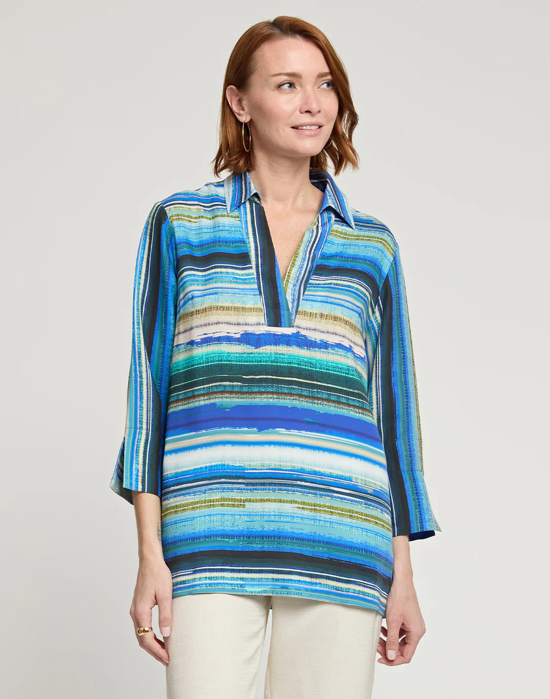 A person with shoulder-length auburn hair is wearing the HINSON WU - VICKY 3/4 Sleeve Tencel Textured Stripe Print Top, which is a loose-fitting shirt featuring vibrant horizontal stripes in blue, green, and white. They are facing slightly left, highlighting the side of the outfit against a plain light gray background.