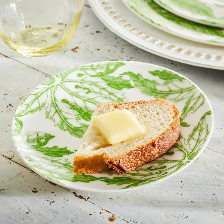 A slice of rustic bread with butter rests on a CASKATA - FREYA PLATE by CASKATA, set on a white wooden table, with a glass and another plate in the background.