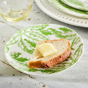 A slice of rustic bread with butter rests on a CASKATA - FREYA PLATE by CASKATA, set on a white wooden table, with a glass and another plate in the background.