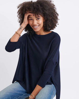 A smiling woman with curly hair wears the MERSEA - CATALINA CREWNECK SWEATER in navy blue and jeans, standing against a plain light background with her hands near her head.
