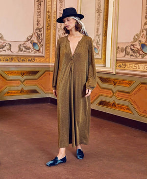 A person stands in a lavishly decorated room, wearing the shimmering long MOMONI - AMEDE DRESS IN LUREX JERSEY, made in Italy by MOMONI, paired with a black wide-brimmed hat adorned with a colorful band. Their hands are in their pockets, and there's a large mirror reflecting part of the ornate wall design.