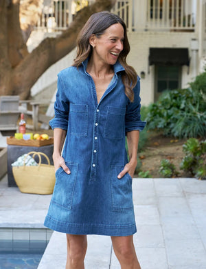A person wearing the FRANK & EILEEN Southern Ireland Utility Dress in Italian Dream Light Denim 1984 Wash, featuring long sleeves, large front pockets, and white buttons, stands outdoors with one hand in their pocket. Their face is not visible. The dress’s classic style subtly nods to practical Irish design.