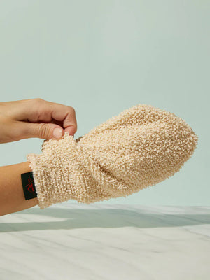 A hand showcases a textured beige KATE MCLEOD - THE BODY MITT against a light turquoise background, while another hand adjusts the cuff with its black tag and red stitching. Beneath, the white surface featuring gray veining creates an ideal exfoliating spa treatment ambiance.