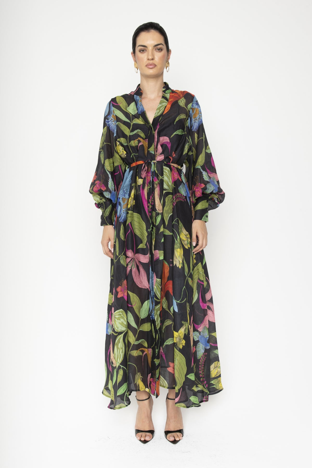 Wearing the LAVI COUTURE - ARTEMISIA FLORAL DRESS, a woman showcases a flowing black dress with vibrant botanical designs in pink, blue, green, and yellow. Against a white background, she highlights its cinched waist and long sleeves while pairing it with black strapped heels.