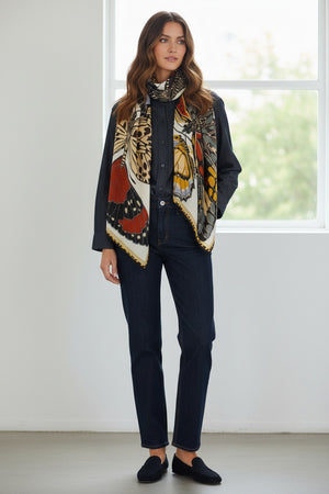 A woman stands facing forward on a plain white background, wearing a black top and pants with the INOUI EDITIONS - PAPILLONS (BUTTERFLIES) WOOL SCARF IN WHITE by INOUI draped around her neck. This beautiful hand-printed scarf, featuring colorful butterfly wing patterns, is crafted in India.